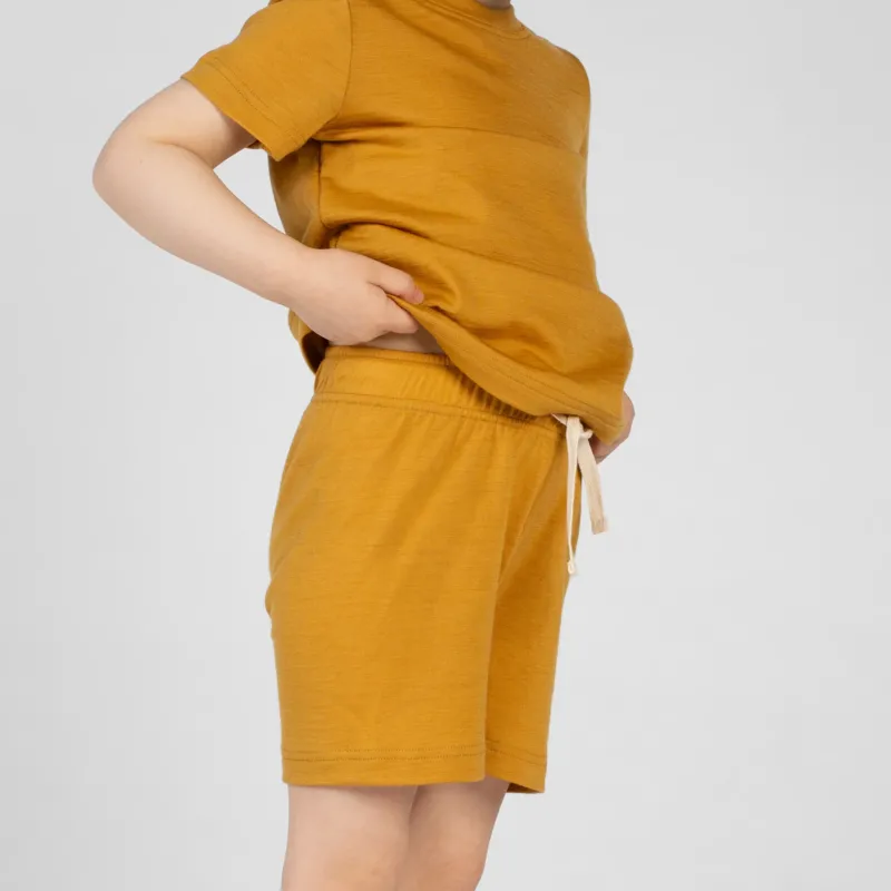A close-up side profile view of a young child's torso and upper legs. The child is wearing a mustard yellow merino short-sleeve t-shirt and matching mustard yellow merino shorts. The child's hand is slightly lifting the hem of the t-shirt to show the elastic waistband and drawstring of the shorts.