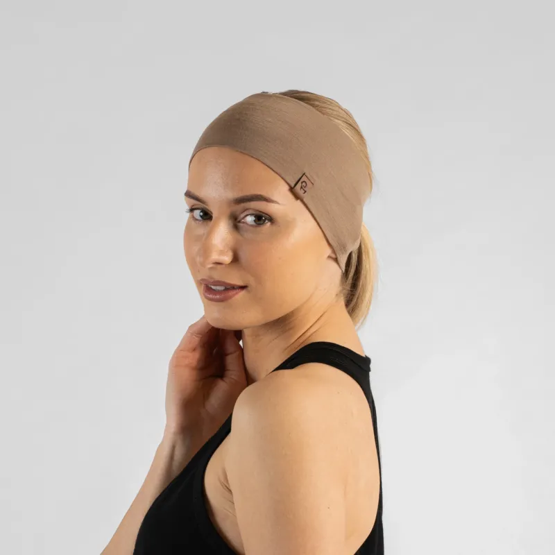 A studio portrait of a woman looking over her shoulder directly at the camera with a gentle smile. She is wearing a wide, light beige knit headband that holds back her blonde ponytail, along with a black tank top. A small, square brown leather logo patch is visible on the side of the headband. The background is a solid light grey.