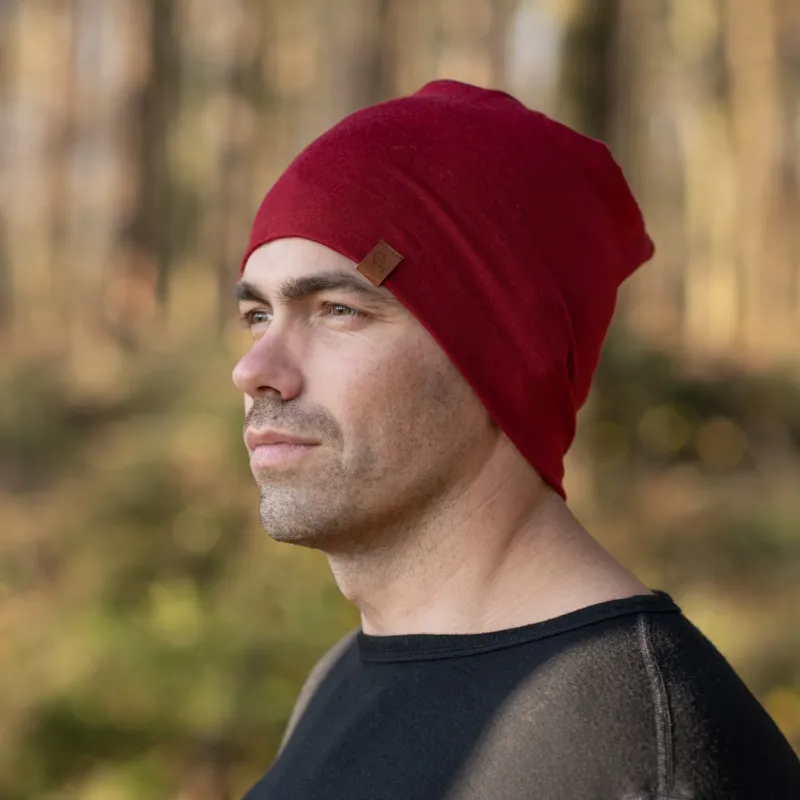 A man standing outside in a forest during fall season, wearing the menique Men's Merino Beanie Hat in Royal Cherry Color.