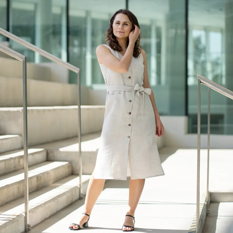 A woman with brown hair, wearing a light beige, sleeveless, button-front dress with a tie belt, poses indoors near concrete stairs.